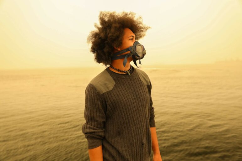 A young man stands in front of a water landscape, his side profile pointed to the right and a gas mask on his face. He wears a dark gray sweater and his hair blows slightly in the wind.