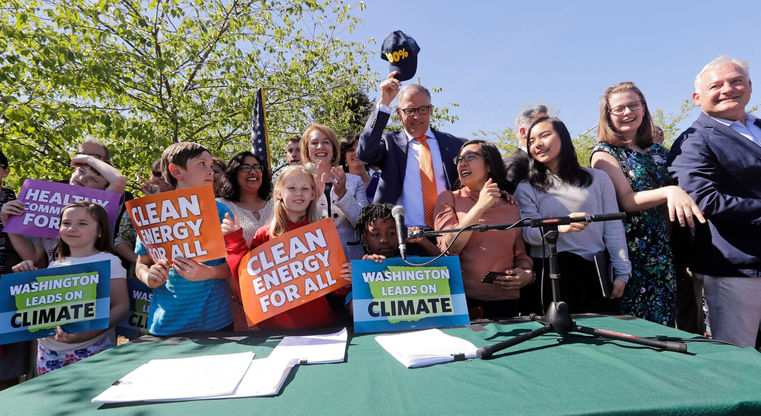 A cluster of children and adults surround a table with a microphone, outdoors. Some hold signs that say "Clean Energy for All" and "Washington Leads on Climate Change." In the center, an older man in a suit holds up a baseball cap.