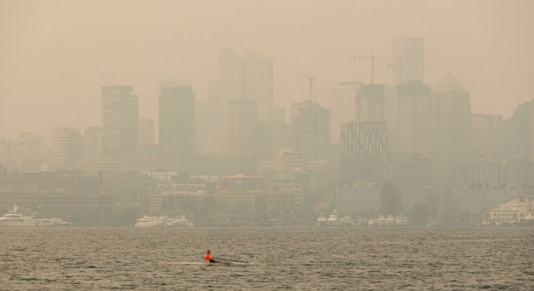 The Seattle skyline, heavily obscured by gray-orange smoke to the point where the buildings are barely visible. At the bottom, Lake Union, where a lone kayaker travels across in a bright orange shirt.