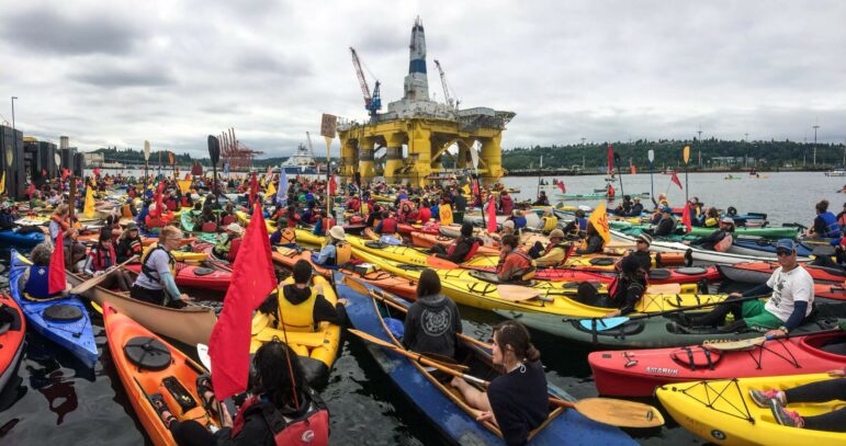 Many kayakers in colorful kayaks crowd together in the water. In the distance is an oil rig.