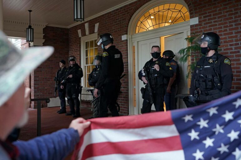 Law enforcement officers with guns stand guard in front of a mansion entryway. In the foreground, arms from off-camera hold up an American flag.