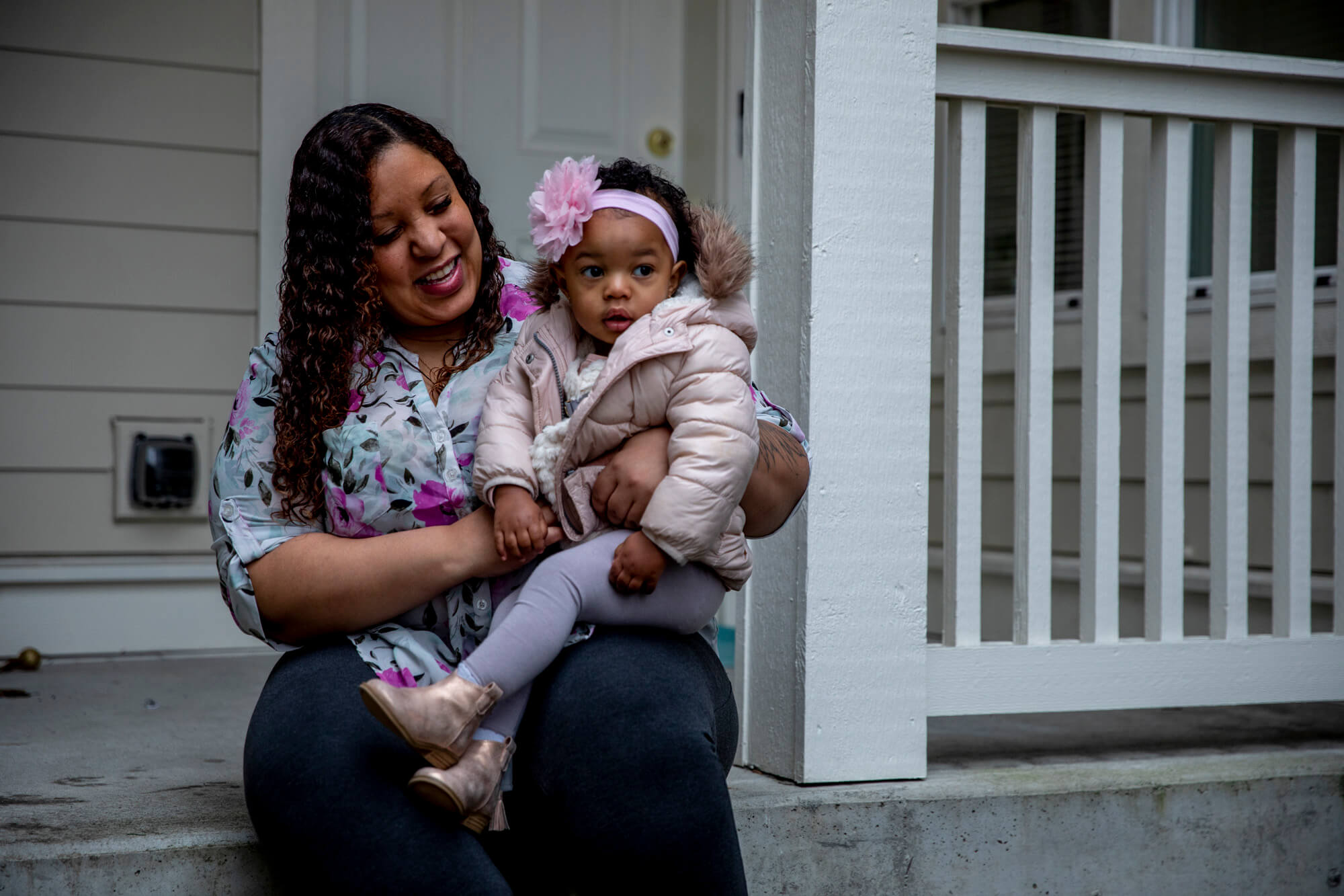 A mother sits on a house porch holding her toddler and smiling.