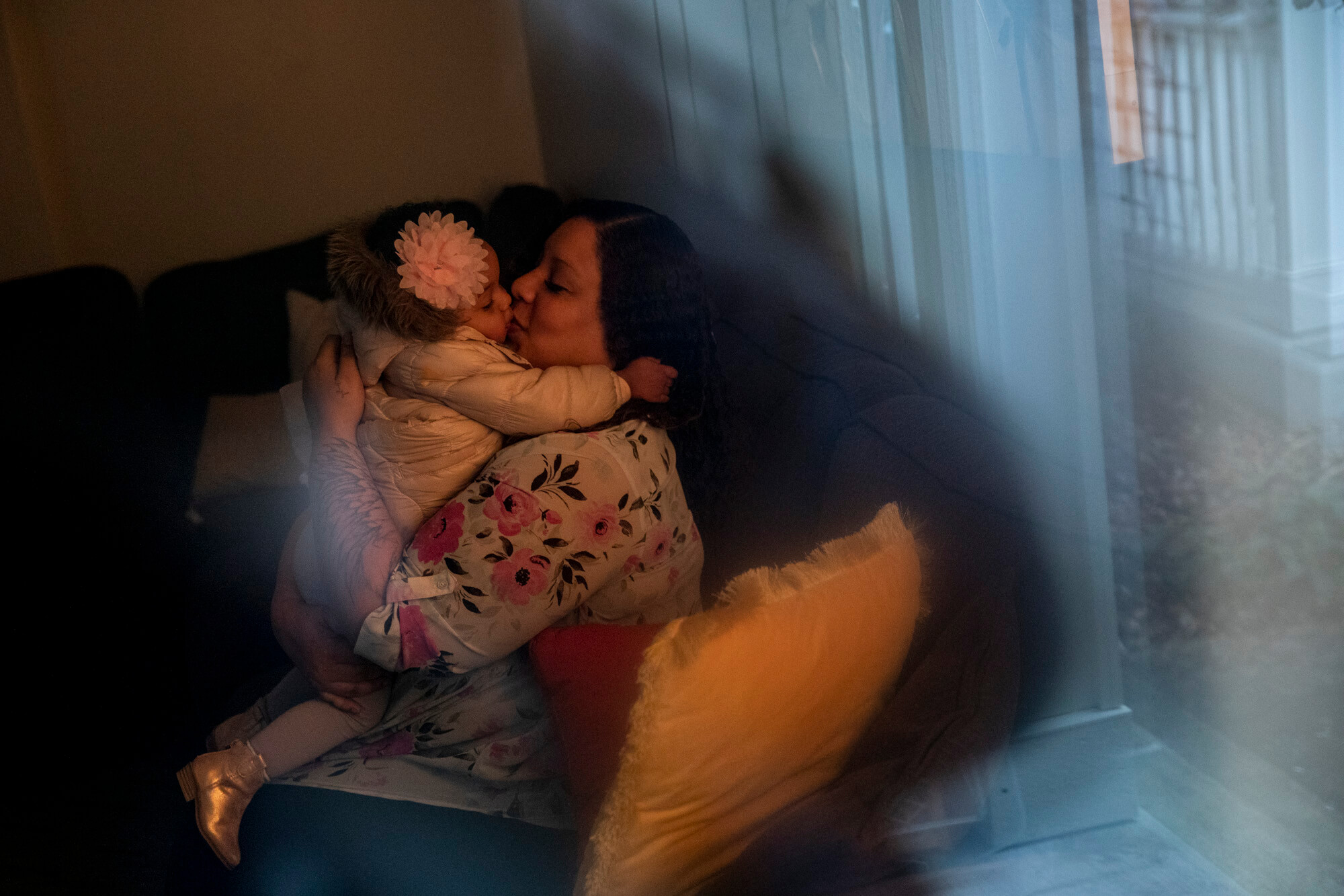 A photo of a mother sitting on a couch indoors, holding her child and kissing her forehead.