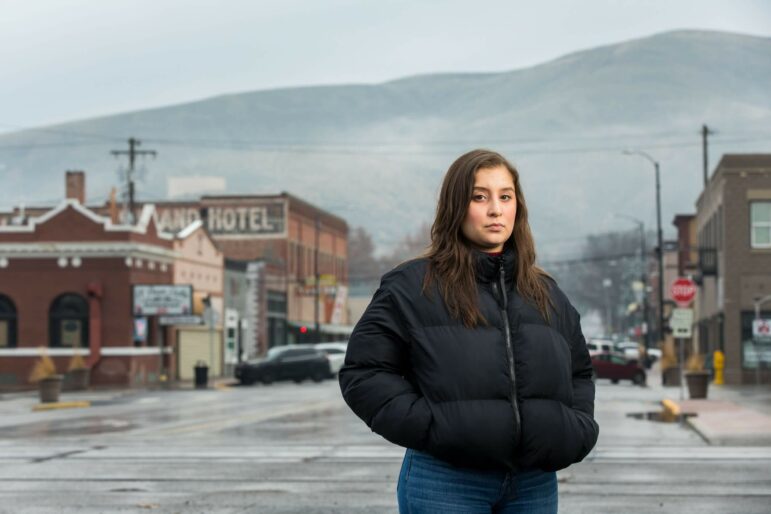 A woman stands in the middle of a street, wearing a winter jacket and looking at the camera.