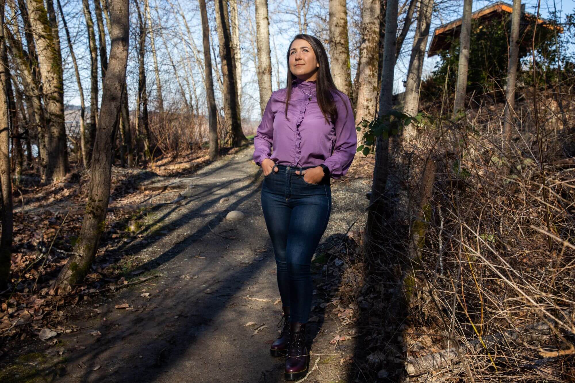 A person stands in the middle of a wooded area, wearing a pink blouse and dark jeans.