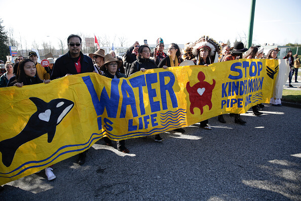 A crowd of protesters carry a yellow banner that says "Waster is life."