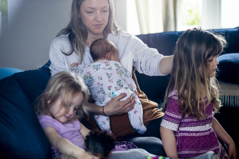 A mother holds a baby while sitting on a couch. Her toddler daughter sits next to her, and a slightly older daughter stands near the couch.