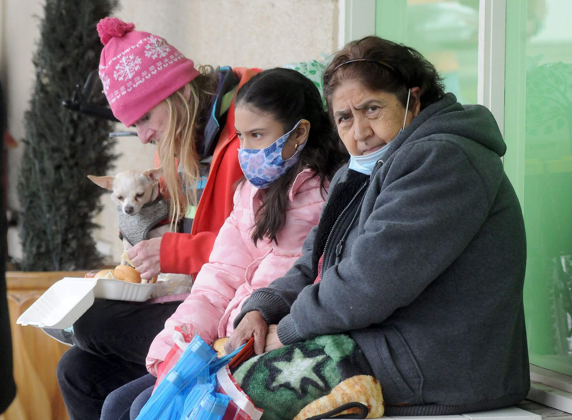 Three people sit on a bench, wearing coats and face masks.
