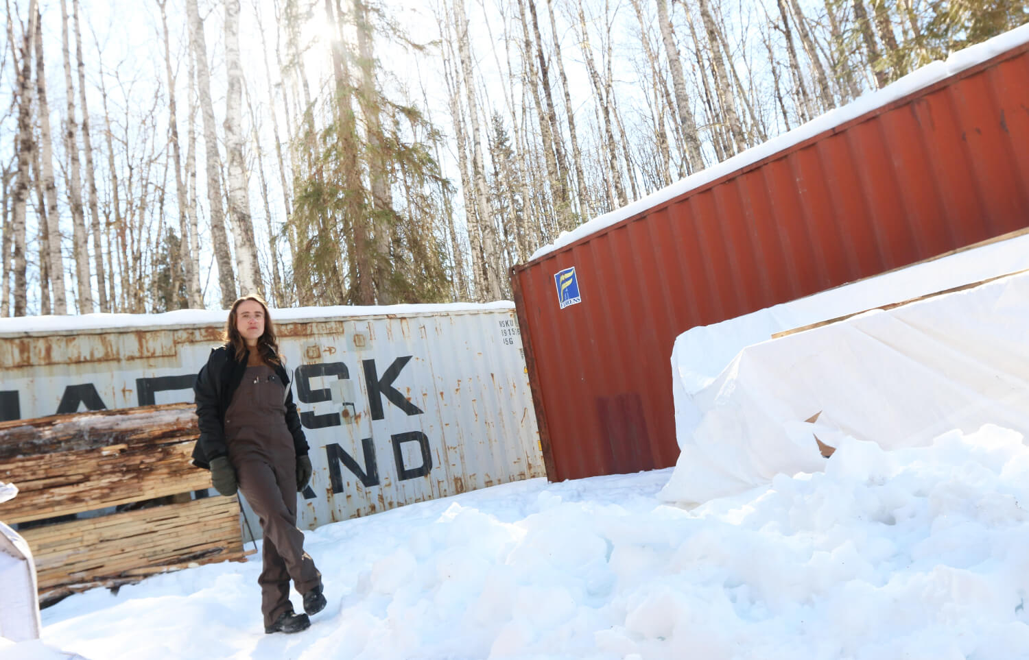 A person standing in the snow next to shipping containers.